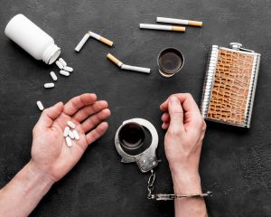Manos agarrando difentes tipos de drogas y con una cadena que representa que tiene una adicción